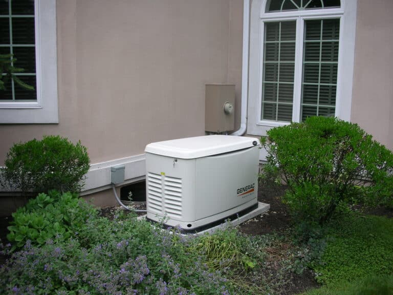 A standby generator is installed on the ground near a house’s exterior wall, surrounded by bushes and plants.