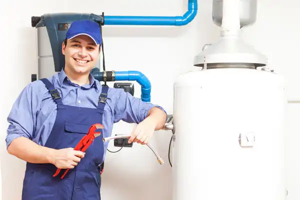 A person in blue overalls and a cap holds plumbing tools while standing next to a water heater with pipes.