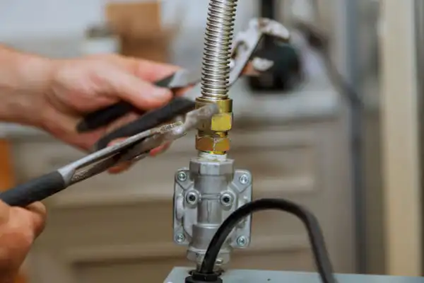 Hands using a wrench to tighten a metal pipe connection on a gas valve. Tools and mechanical components are visible in the setup.