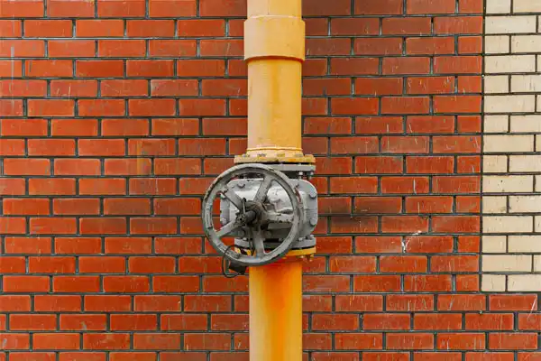 A yellow pipe with a metal valve is mounted on a brick wall featuring red and beige bricks.
