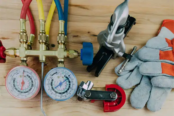 A set of HVAC tools on a wooden surface, including gauges with colored hoses, a pipe cutter, a clamping tool, and a pair of work gloves.