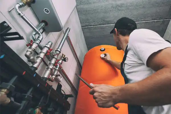 Technician in overalls adjusts valves on orange boiler tank in utility room.