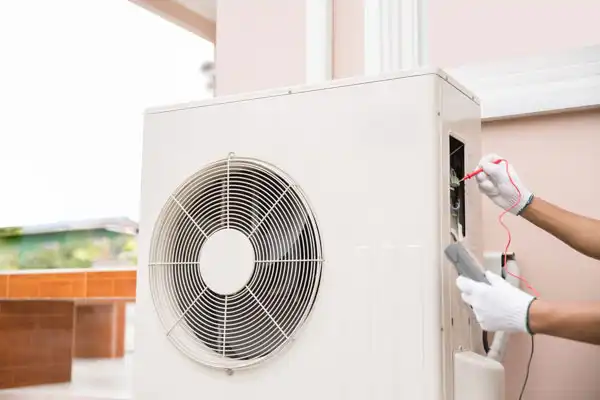 Technician using a multimeter to check an air conditioning unit outdoors.