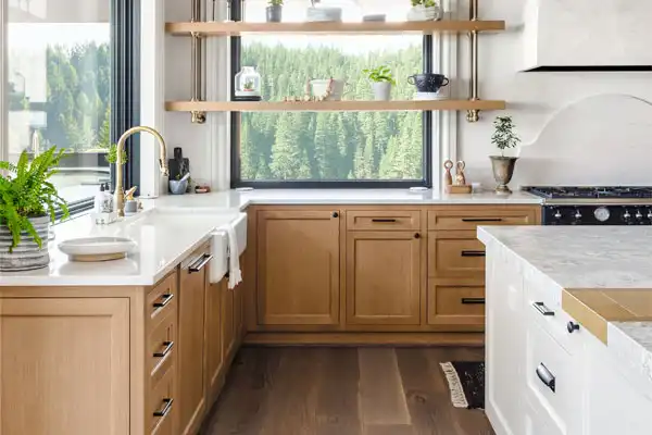 Modern kitchen with light wood cabinets, white countertops, and black appliances. Shelves hold plants and dishes. Large window offers a view of green trees.