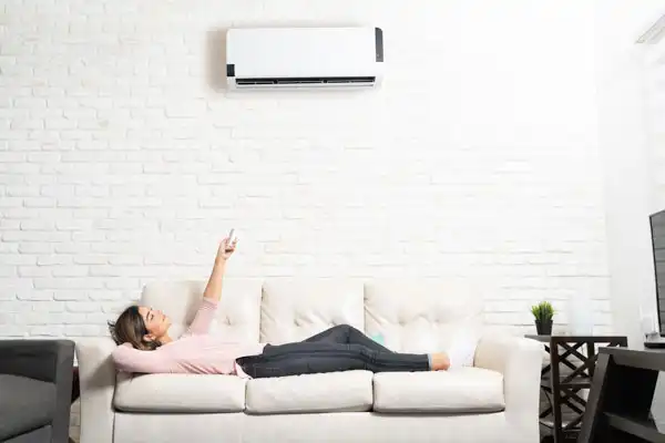 Person lying on a couch in a white room, holding a remote control and pointing it towards an air conditioner mounted on a brick wall.