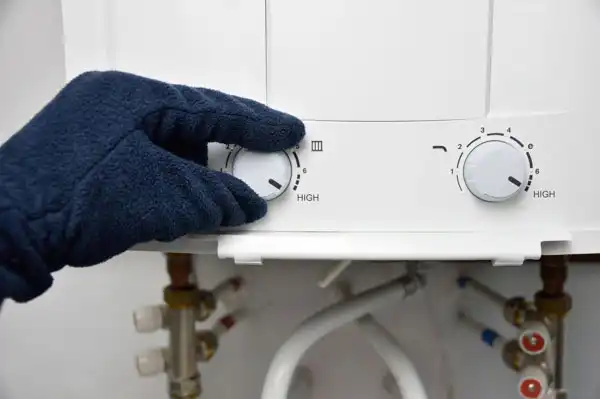 A gloved hand adjusts the temperature dial on a water heater, with visible pipes and connections below.