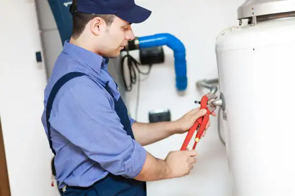 A person in a blue cap and uniform uses a wrench to adjust a valve on a large cylindrical tank in a utility area.