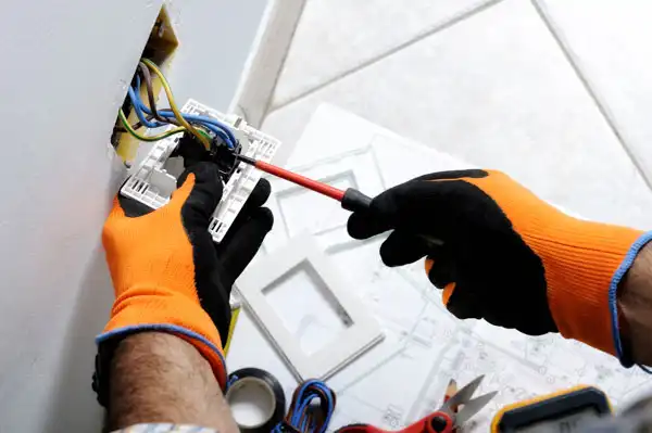 Person wearing orange gloves uses a red screwdriver to install or repair a wall electrical outlet, with wires exposed.