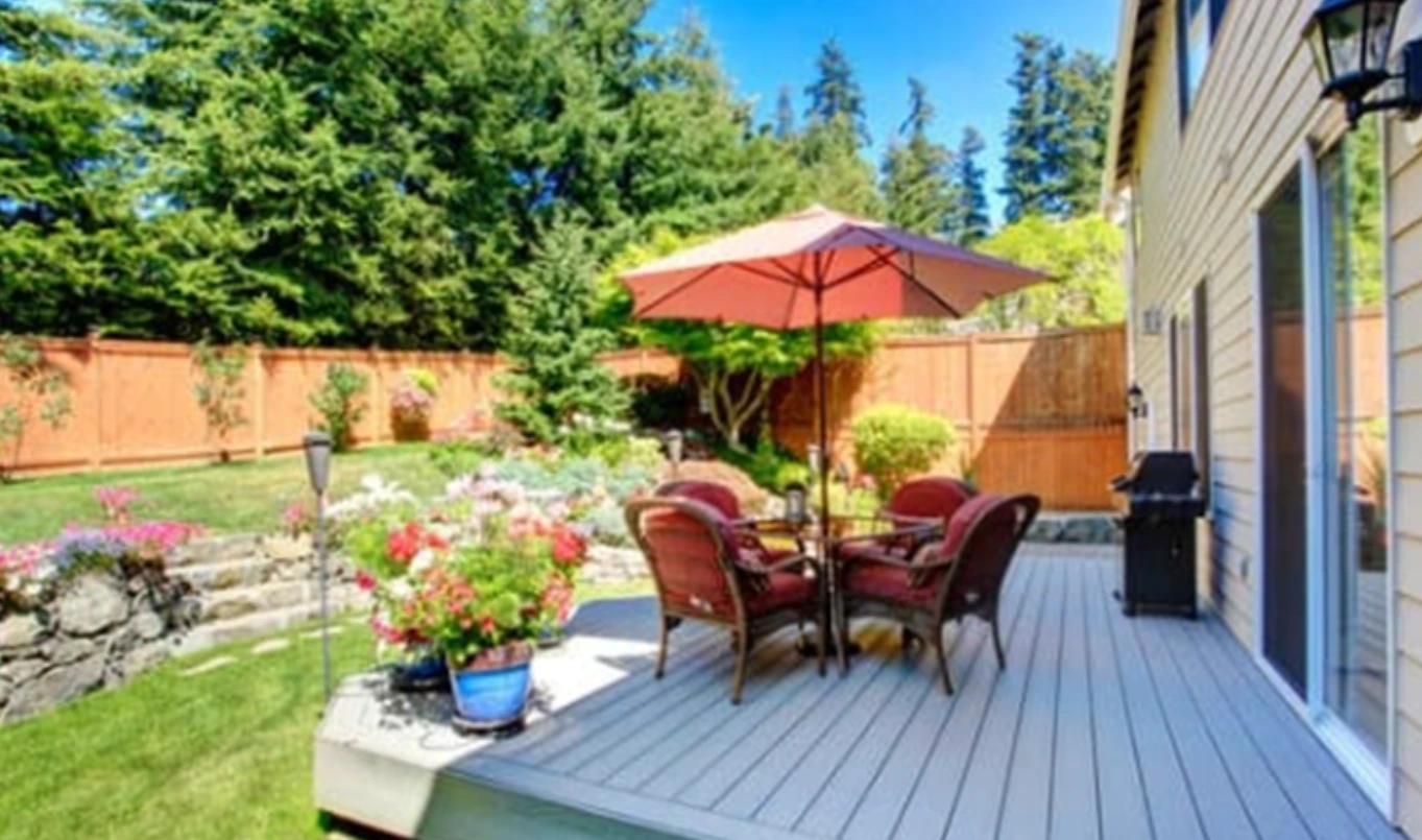 A backyard deck with a round table, four chairs, an umbrella, potted plants, a grill, and a wooden fence surrounded by trees and greenery.