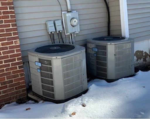 Two outdoor air conditioning units are installed on the ground next to the side of a house, surrounded by snow.