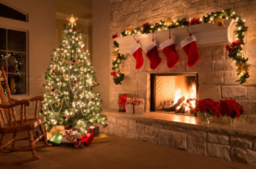 A decorated Christmas tree with presents stands next to a lit fireplace, which is adorned with four red stockings and festive garland in a cozy living room.