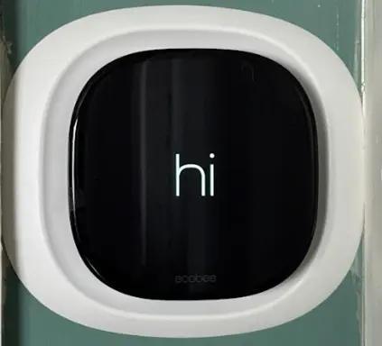 A smart thermostat with a black screen displays the word "hi" in white text, set in a white frame on a green wall.