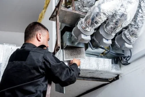 A technician in a black uniform services an HVAC system, inspecting and working on ductwork and insulated pipes attached to the ceiling.