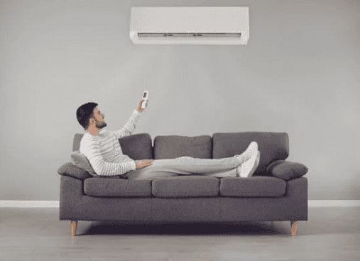A man lies on a gray sofa using a remote control to operate an air conditioner mounted on a gray wall.
