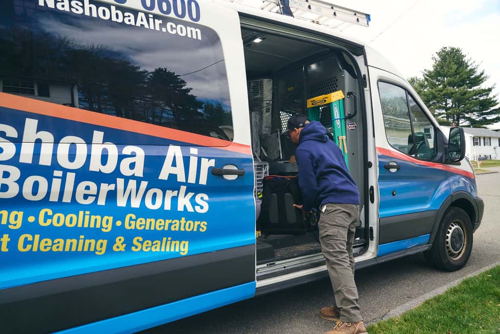 A worker in a blue hoodie and cap stands at the open side door of a Nashoba Air & BoilerWorks service van, reaching inside for tools or equipment.