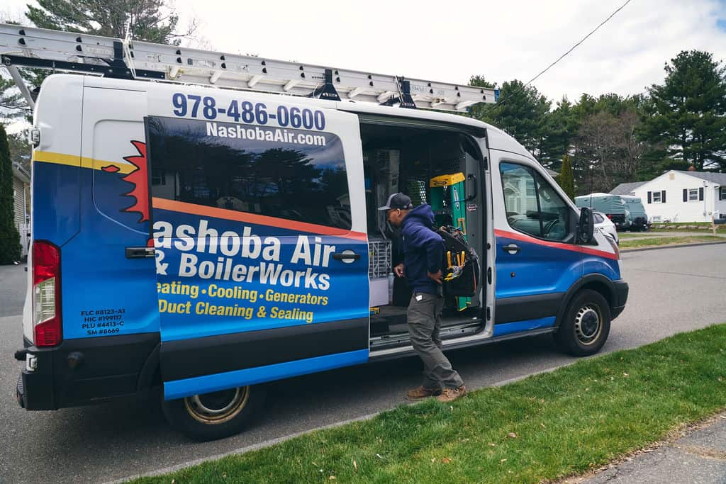 A technician stands beside an open Nashoba Air & BoilerWorks van, preparing equipment for a job in a residential neighborhood.