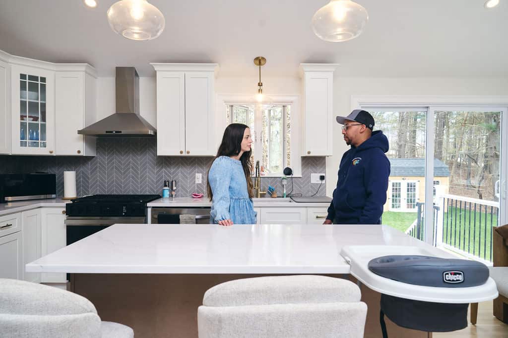Two people stand and talk across a white kitchen island in a modern kitchen, with windows looking out onto a green backyard.