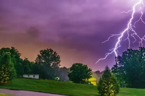 A bolt of lightning strikes near a grassy field and trees under a purple, stormy sky at dusk.