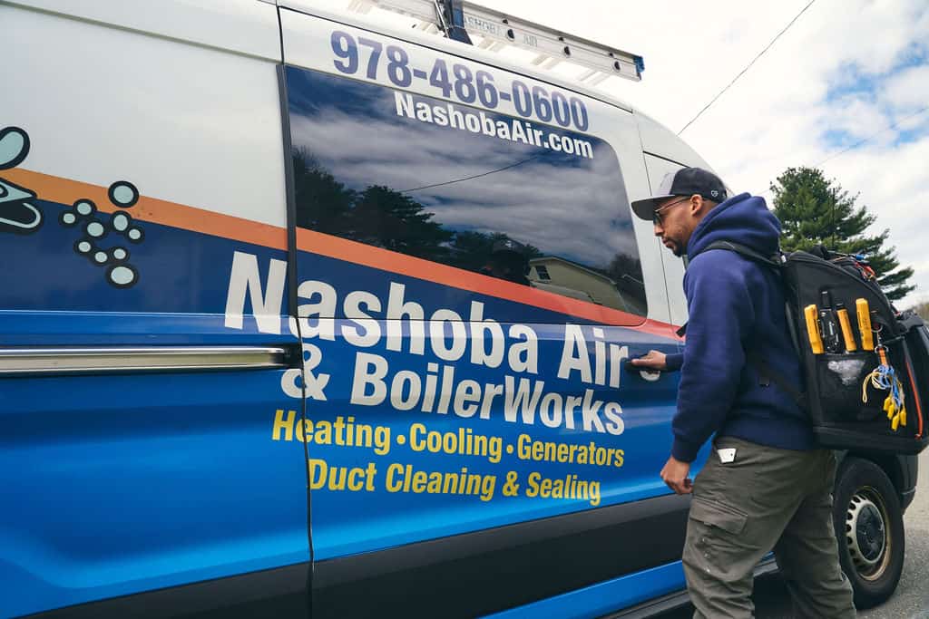 A person wearing a backpack with tools opens the door of a Nashoba Air & BoilerWorks service van, which advertises HVAC, AC Repair & Cooling Services, and duct services.