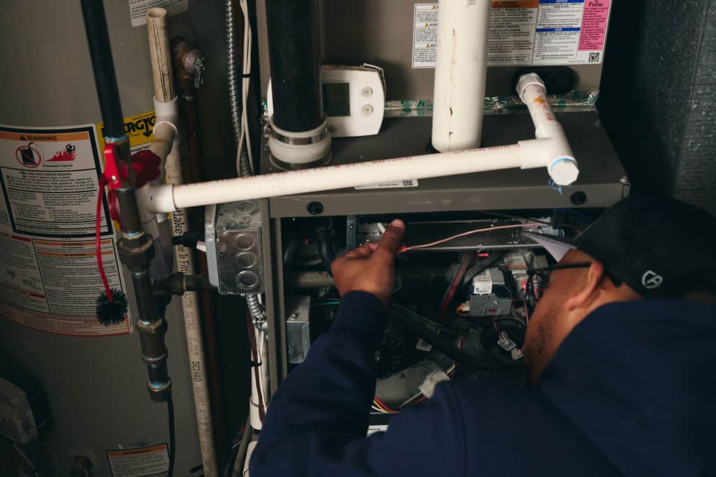 A person in a blue hoodie inspects and repairs the interior of a furnace next to a water heater, with various pipes and wires visible—demonstrating expertise often associated with AC Repair & Cooling Services.