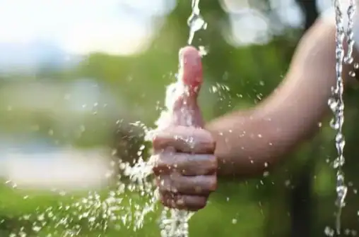 A hand showing a thumbs-up gesture is being splashed with running water outdoors, with a blurred green background.